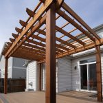 Wooden pergola over a spacious deck attached to a house, with brown beams and clear blue sky above.