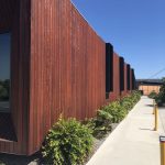 Brown vertical wood-clad wall with black rectangular fins along a sunny sidewalk.