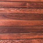 Close-up of horizontal red-brown wooden planks with small metal screws along the seams, showing wood grain and natural imperfections.