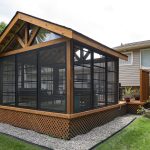 Wood-framed screened porch attached to a beige house, surrounded by green lawn and gravel edging in a backyard.