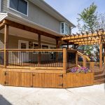 Wooden two-tier deck around a beige house, with a wooden pergola and black metal railing, bordered by flower beds and a concrete patio.