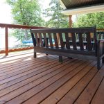 Backyard wooden deck with black outdoor furniture and railing, surrounded by green trees and a sunny sky