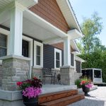 Front porch of a house with stone foundation, white columns, dark siding, and wooden steps; pink flowers in pots.
