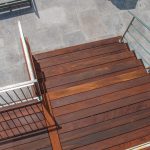 Bird's-eye view of a wooden deck with metal railing leading to a gray stone patio and a flowering garden bed nearby.