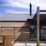 Exterior shot of a modern building with vertical wooden siding, a corrugated metal roof, a black chimney, and a glass door entry under a wooden post structure