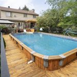 Backyard with a rectangular in-ground pool surrounded by a wooden deck, black fence, and a two-story beige house with trees in the background.