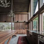 Open kitchen with a long wooden island, dark wood cabinets, antler chandeliers, and large windows showing trees outside.
