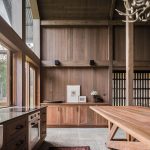 Rustic kitchen interior with warm wood panels, large windows, and a wooden dining table along a counter.