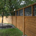 Wooden privacy fence with dark decorative metal panels beside a tree with red berries in a landscaped yard.