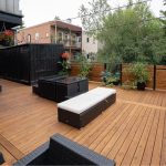 Rooftop wooden deck with black wicker lounge set and white cushions, surrounded by potted plants and a wooden privacy fence.
