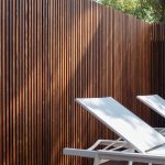 Tall vertical wooden slat fence in a sunny outdoor space, with white lounge chairs visible at the bottom right.