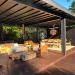 Outdoor lounge area under a dark wooden pergola with wicker sofa, cushions, and a hammock hanging nearby.