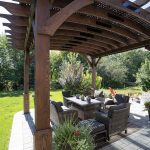 Outdoor dining area under a wooden pergola with wicker chairs and a table on a stone patio in a sunny garden.