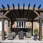 Wooden pergola over a patio with wicker seating in a brick house backyard.