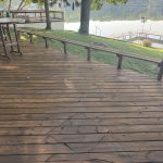 Wooden deck beside a calm lake with a round table, benches, and a dock extending into the water.