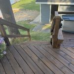Wooden outdoor stairs leading down to a grassy yard, with a gray shed and a grey trash bin nearby; a boat dock and water visible in the background.