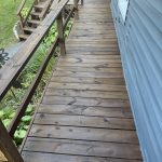 Long wooden deck along the side of a blue house, with a railing and steps leading down to a garden. 2010s style weathered wood.